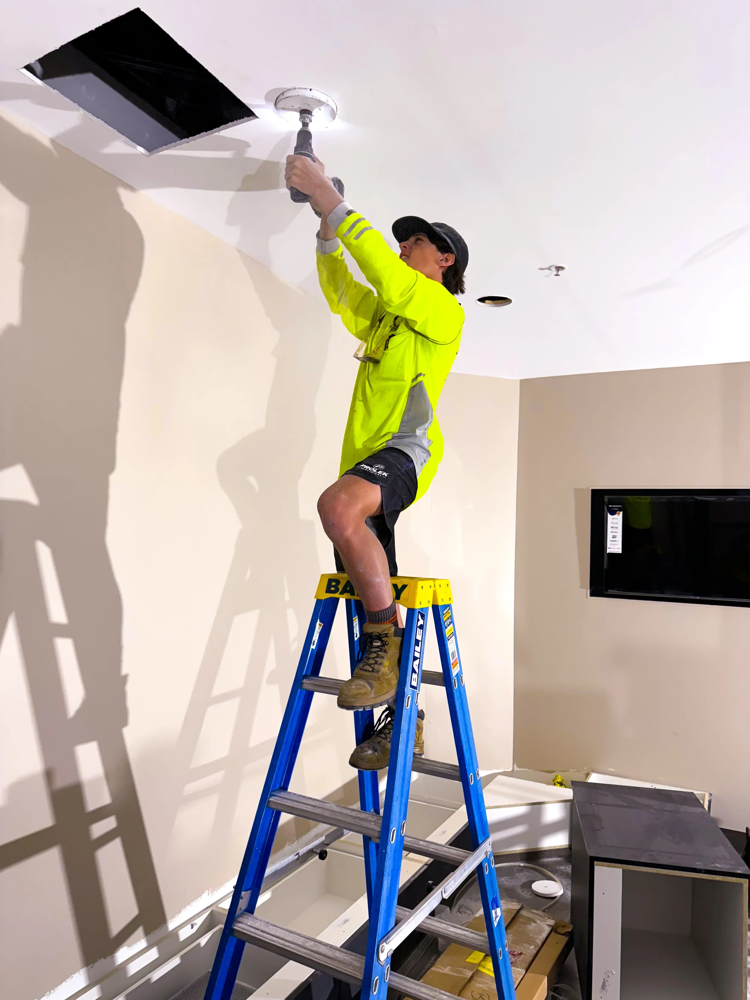 A person standing on a step ladder installing a ceiling vent or light fixture in a room under construction or renovation.