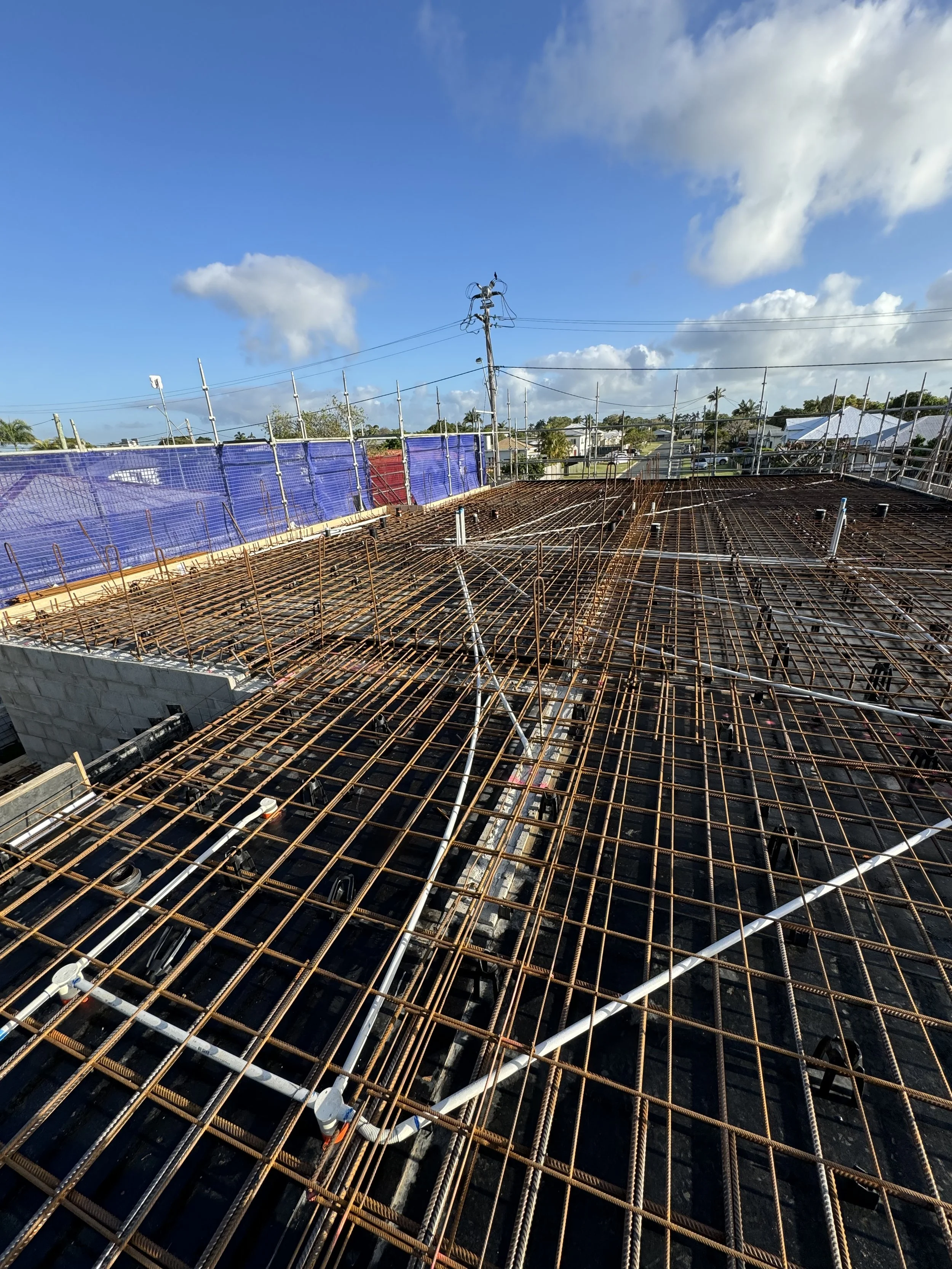 Construction site with rebar grid and blue safety fencing under a partly cloudy sky.