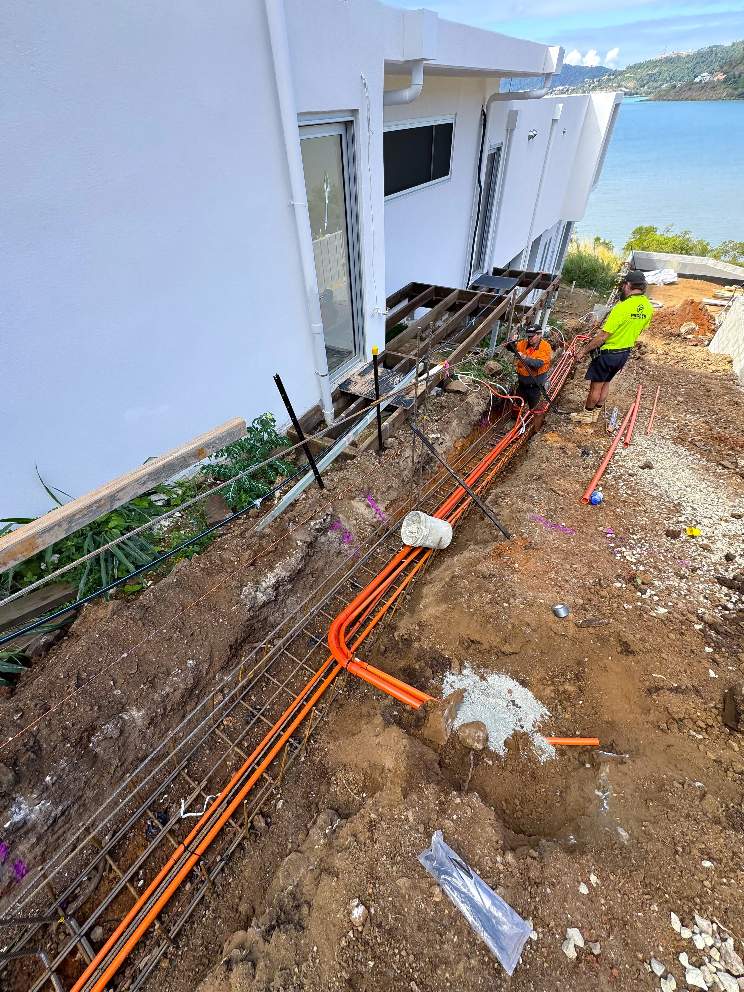 Workers installing electrical or plumbing pipes outside a modern white building overlooking a body of water, with construction materials and dug trench in the foreground.