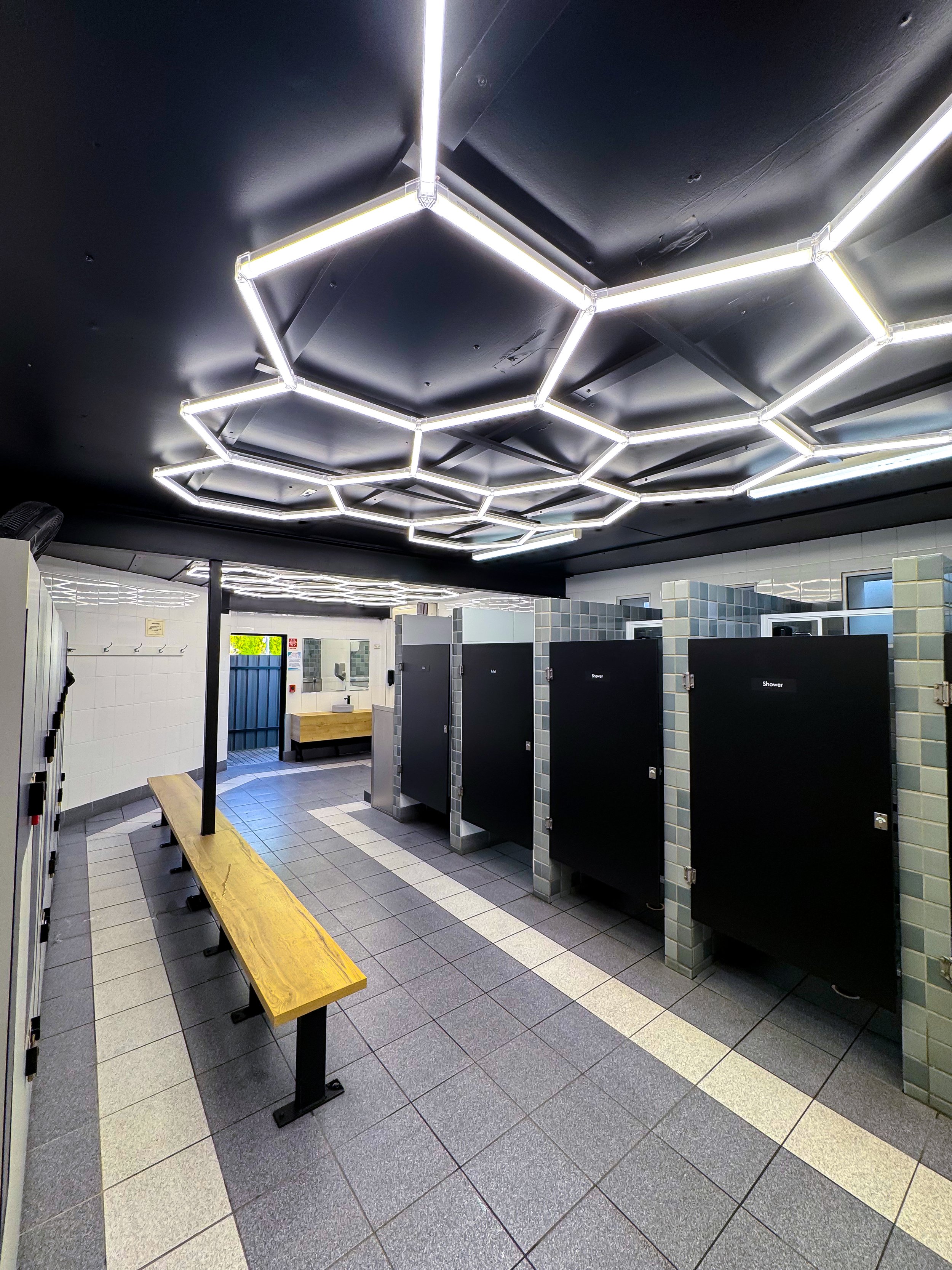 Public shower area with black stall doors labeled "Shower", a wooden bench, tiled walls, and ceiling with modern hexagonal lighting fixtures.