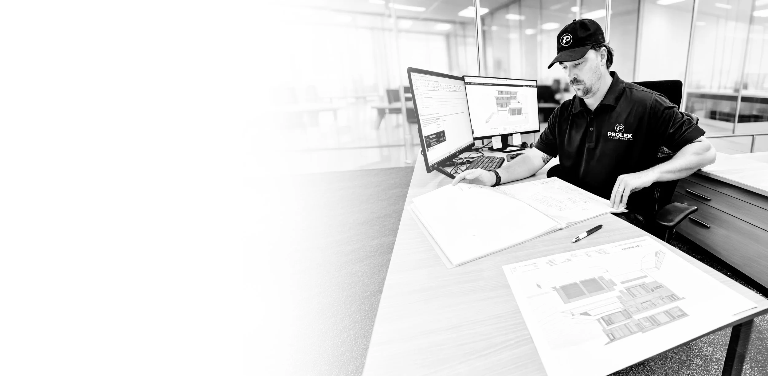 A man working at a desk with two computer monitors, architectural blueprints, and a pen in an office setting.
