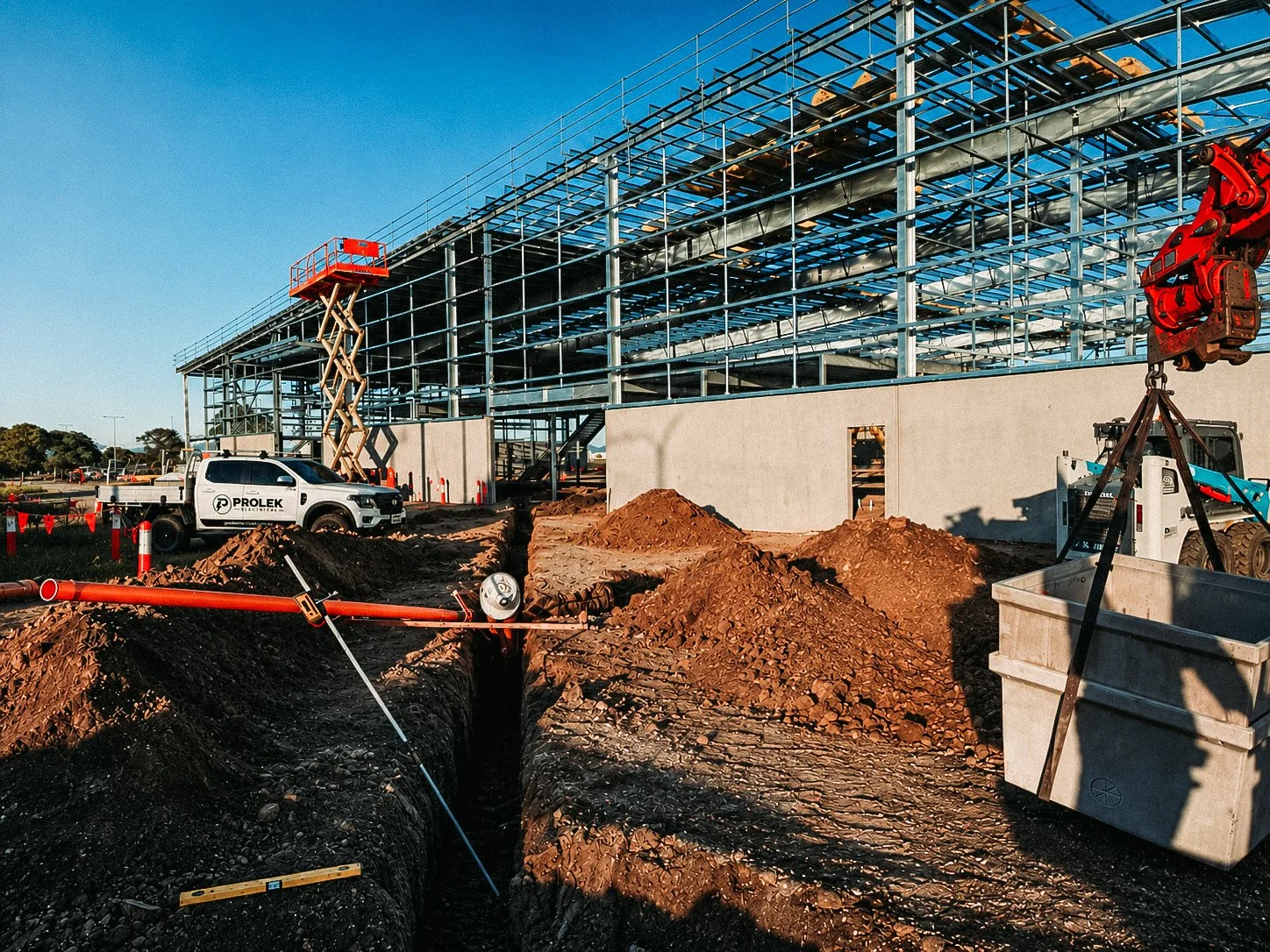 Construction site with steel framework of a building, dirt piles, construction vehicles, a scissor lift, and a crane in a clear weather day.