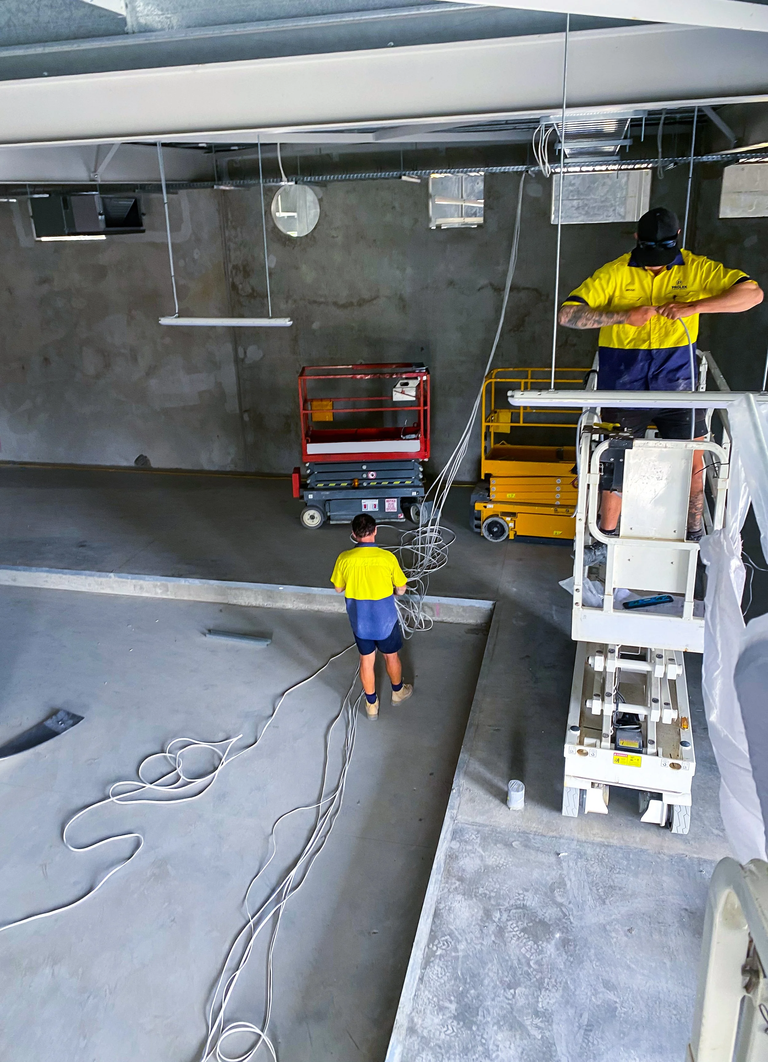 Construction workers installing electrical cables inside a building. One worker is on a lift, while another is on the ground managing cables. Various tools and equipment are visible around them.