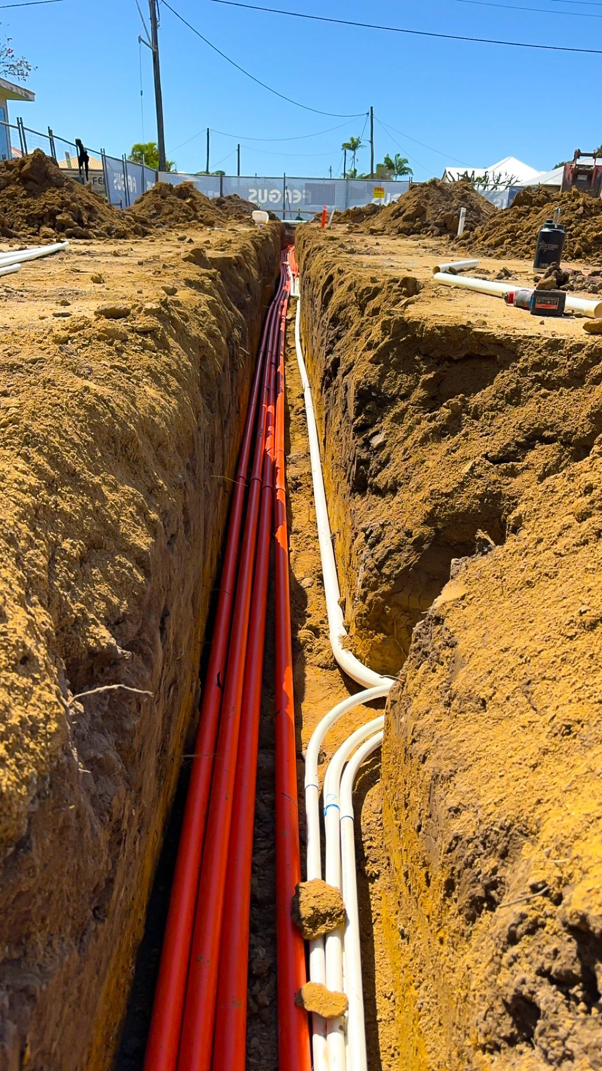 Construction site trench with red and white pipes installed underground, dirt piles on both sides, and blue sky overhead.