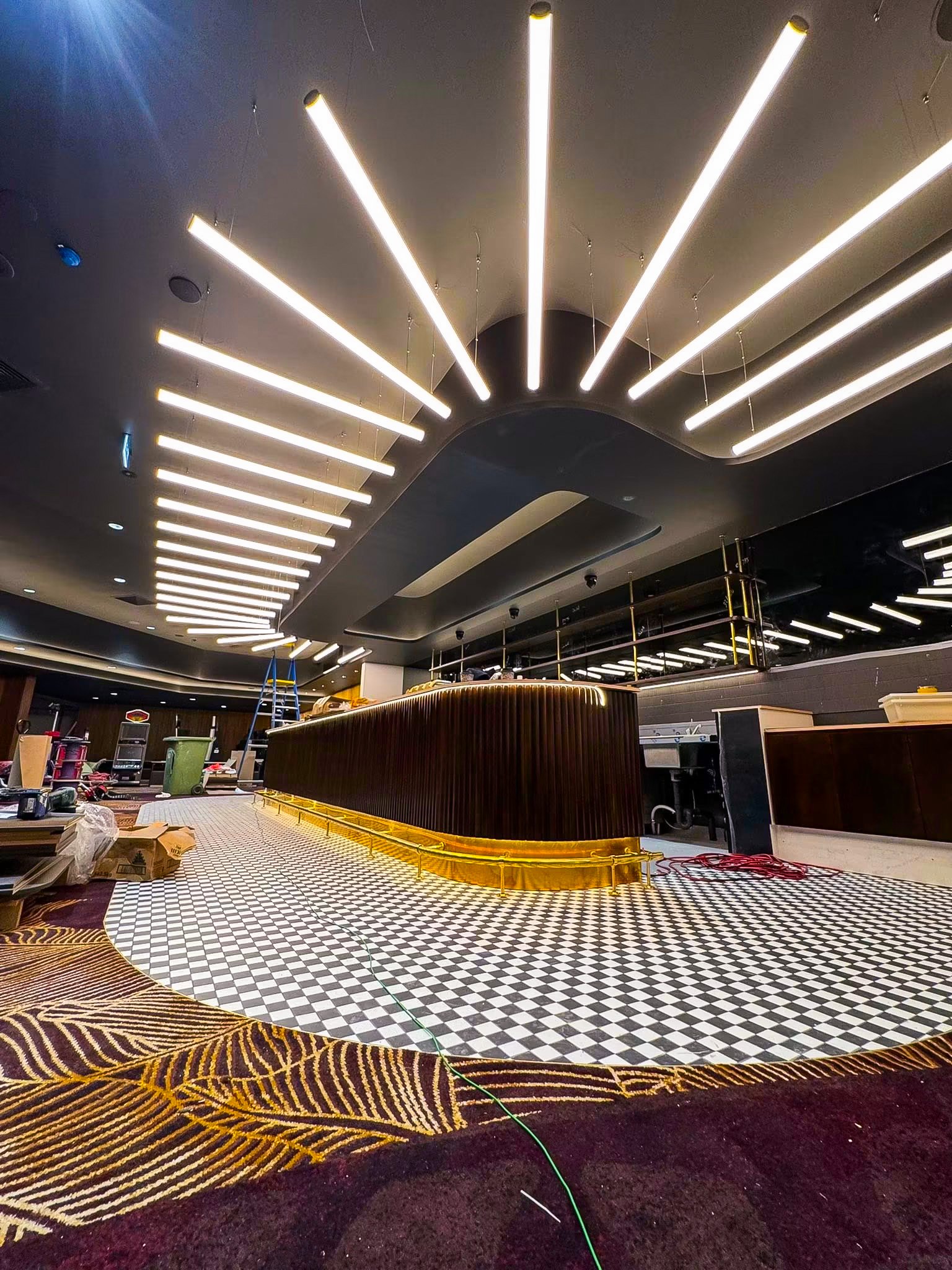 Interior of a space under construction or renovation, featuring a black ceiling with modern LED strip lighting, a checkerboard-patterned floor, and a curved wooden bar with gold trim. Construction materials and tools are visible on the floor.