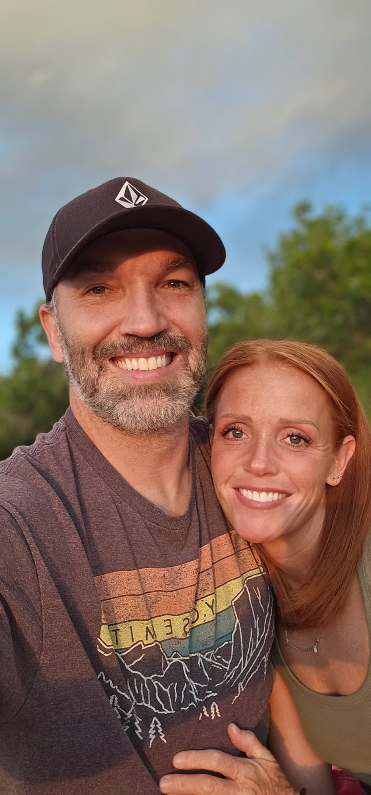 A smiling man wearing a black cap and a woman with red hair smiling, outdoors with a blue sky and green trees in the background.