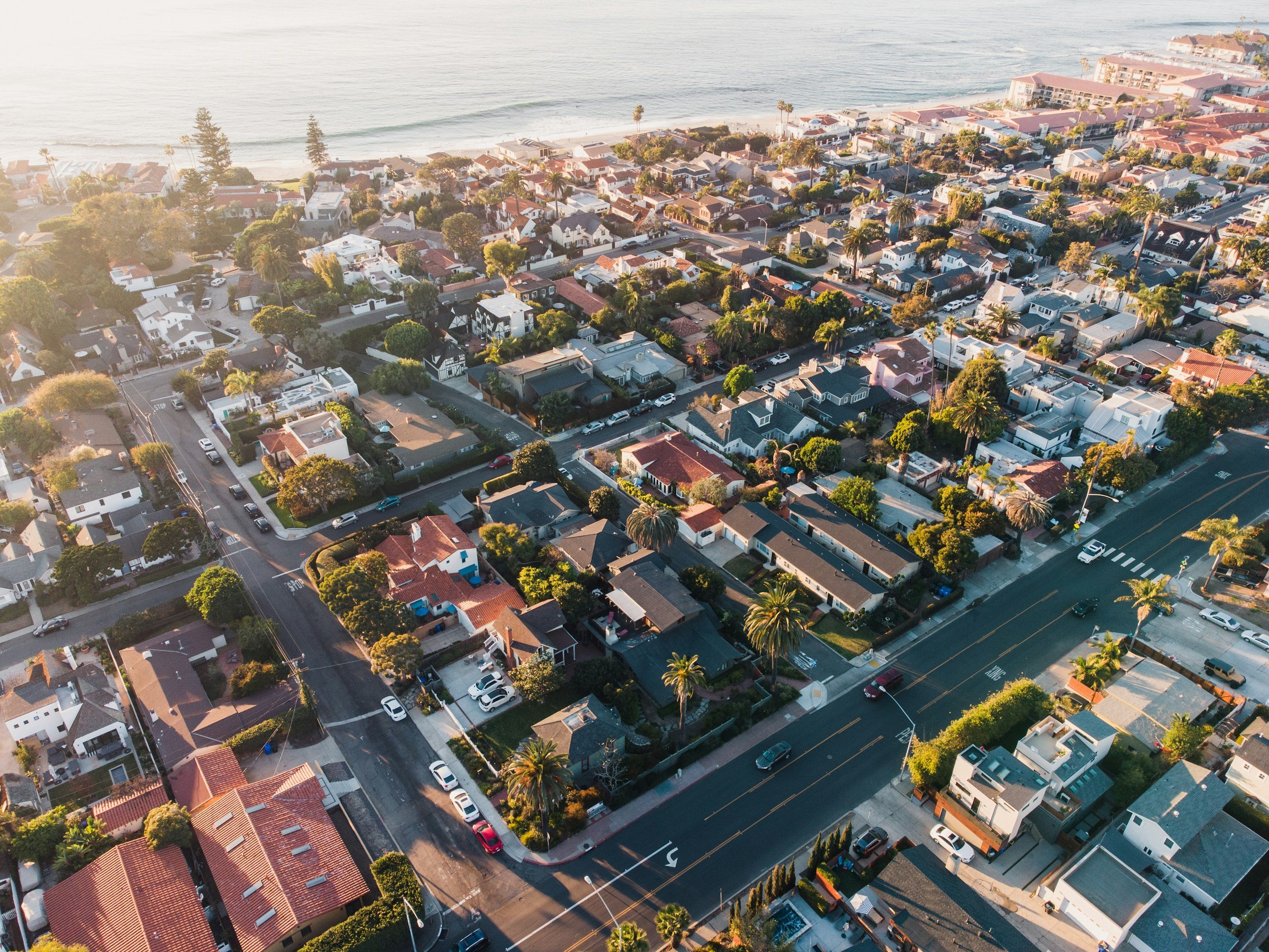 Aerial view of a coastal residential neighborhood with houses, trees, and parking, near the beach and ocean.
