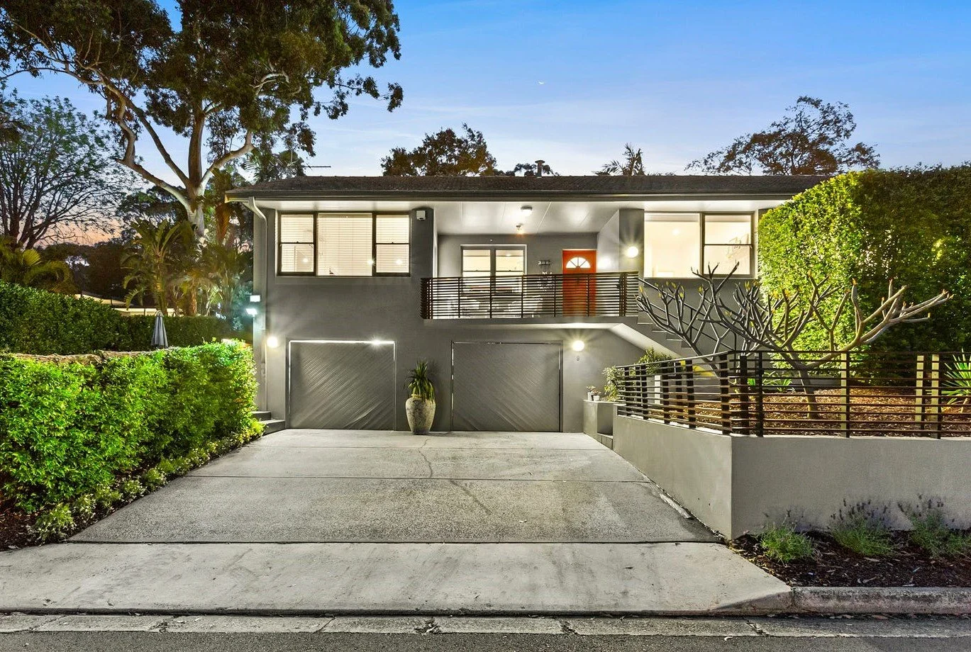 Modern gray two-story house with front driveway, potted plant, and illuminated exterior, during dusk.