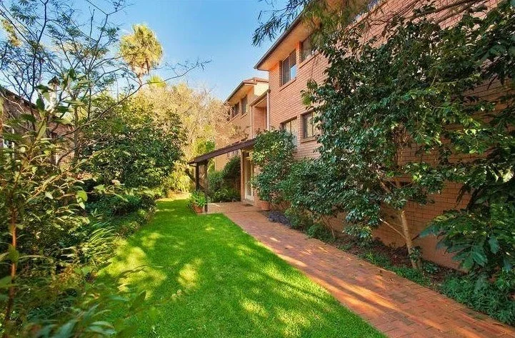 Backyard with a brick pathway and a lush green lawn next to a multi-story brick house surrounded by trees and bushes under a clear blue sky.