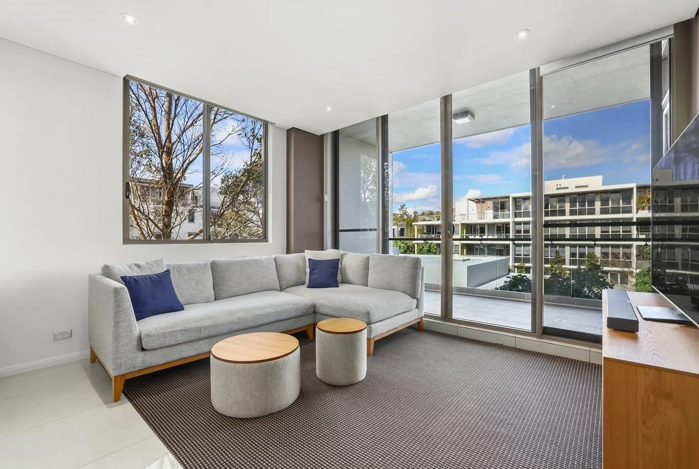 Living room with large window and sliding glass door, light gray sectional sofa with blue pillows, two small round tables, a flat-screen TV, and modern decor.