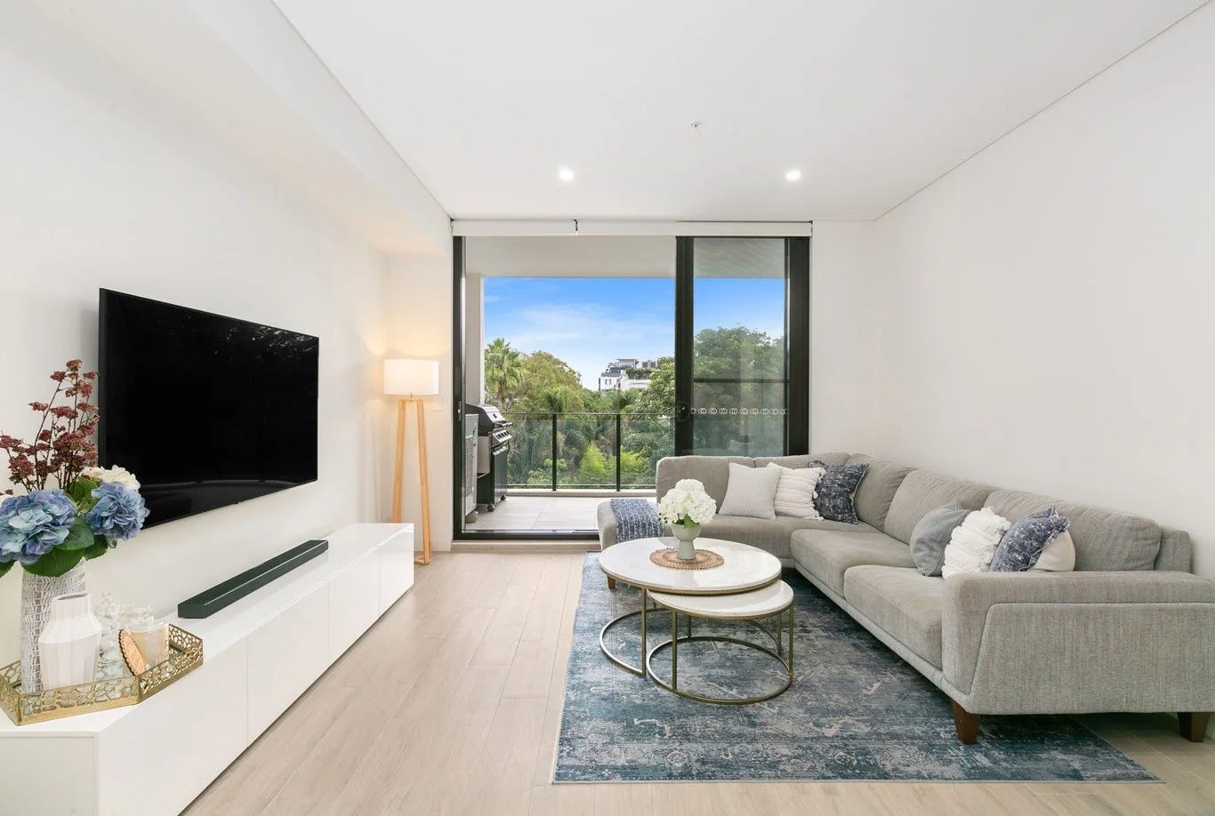 Modern living room with a gray sectional sofa, white cushions, a blue area rug, and a glass sliding door leading to a balcony with trees in the background.