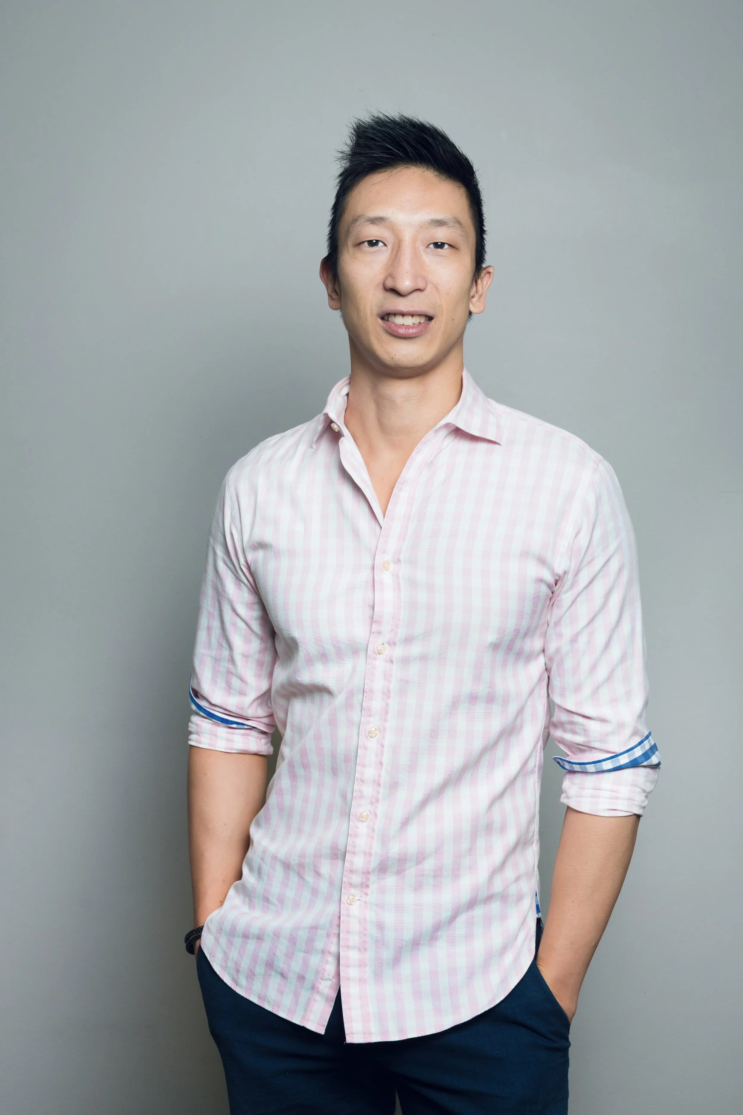 Young man with short black hair, wearing a light pink striped button-up shirt with rolled sleeves and dark pants, standing against a plain gray background with one hand in his pocket.