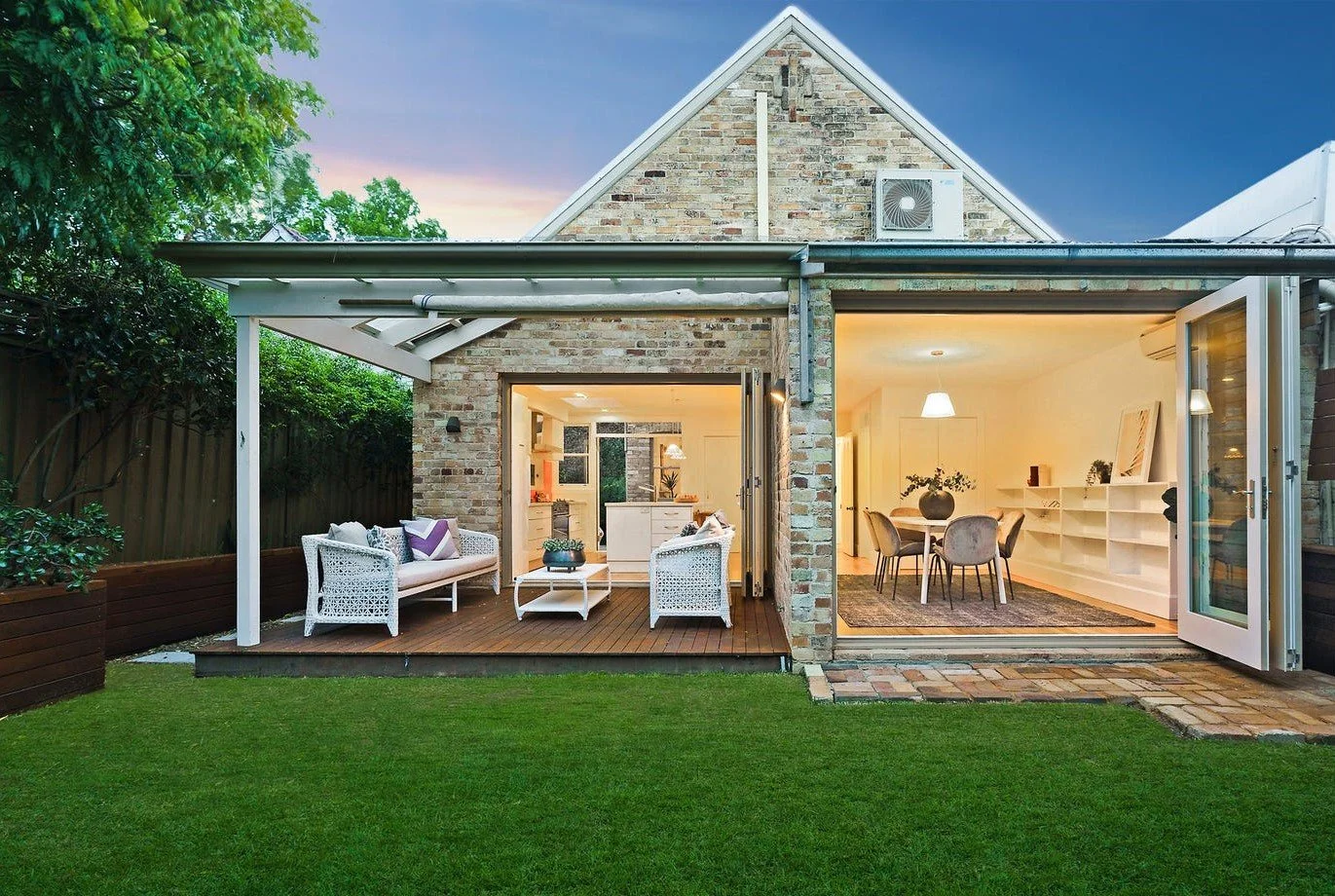 Backyard view of a brick house with a living room and dining area visible through large open glass doors, outdoor seating on a wooden deck, green lawn, and trees at dusk.