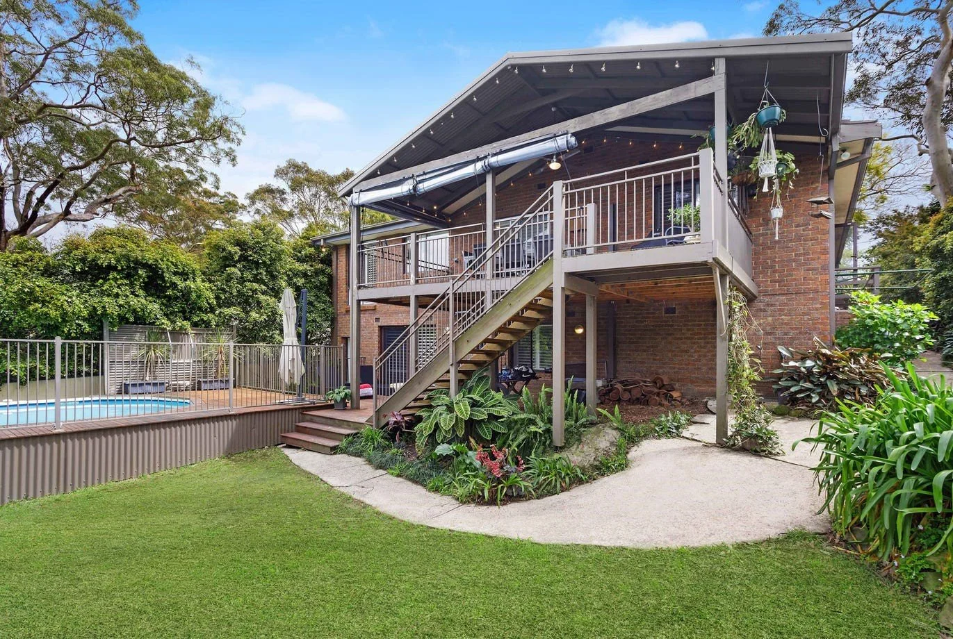 Backyard view of a brick house with a raised deck, outdoor staircase, and a swimming pool enclosed by a safety fence.