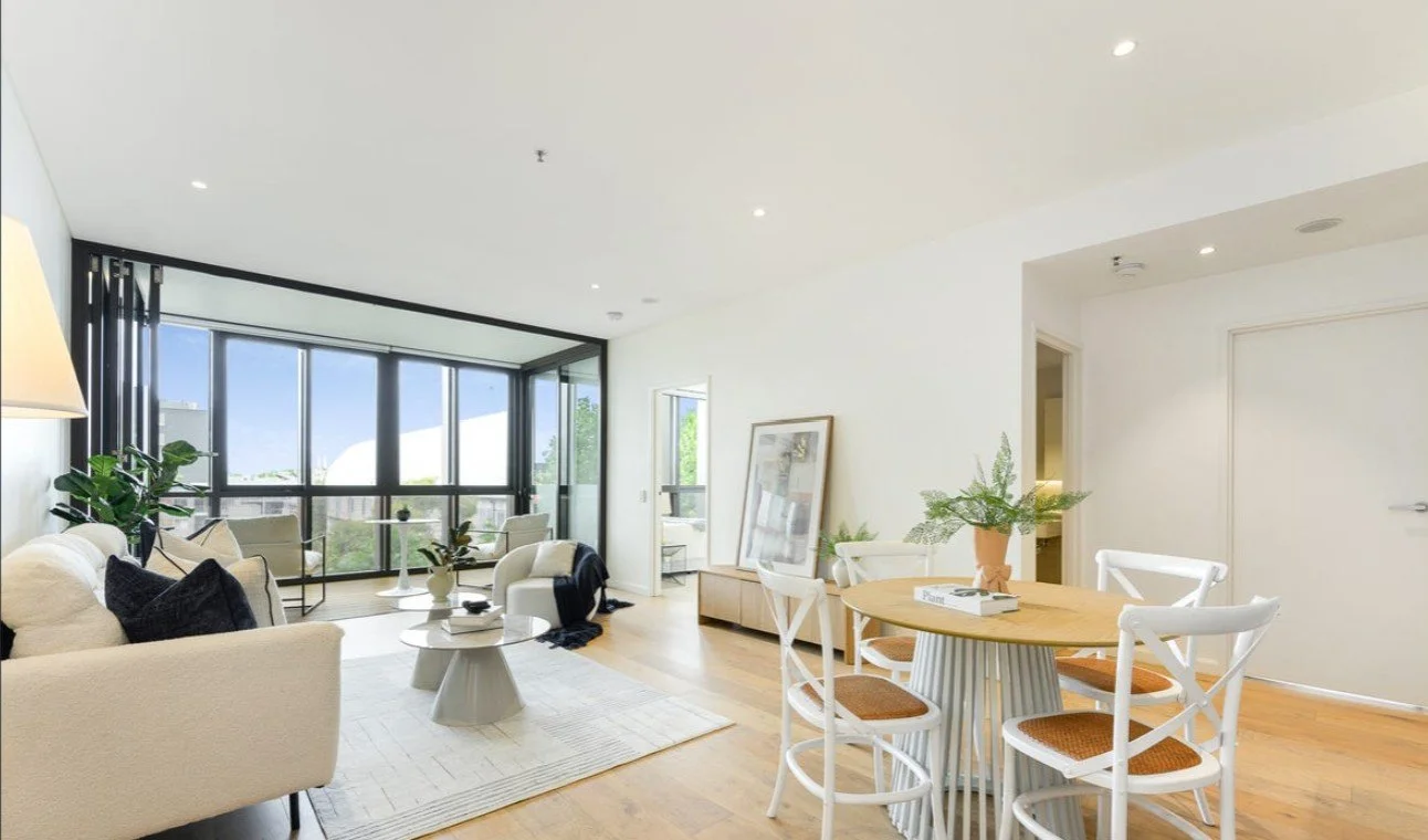 Bright living room with large windows, white sofas, a round wooden dining table with four chairs, and various potted plants.