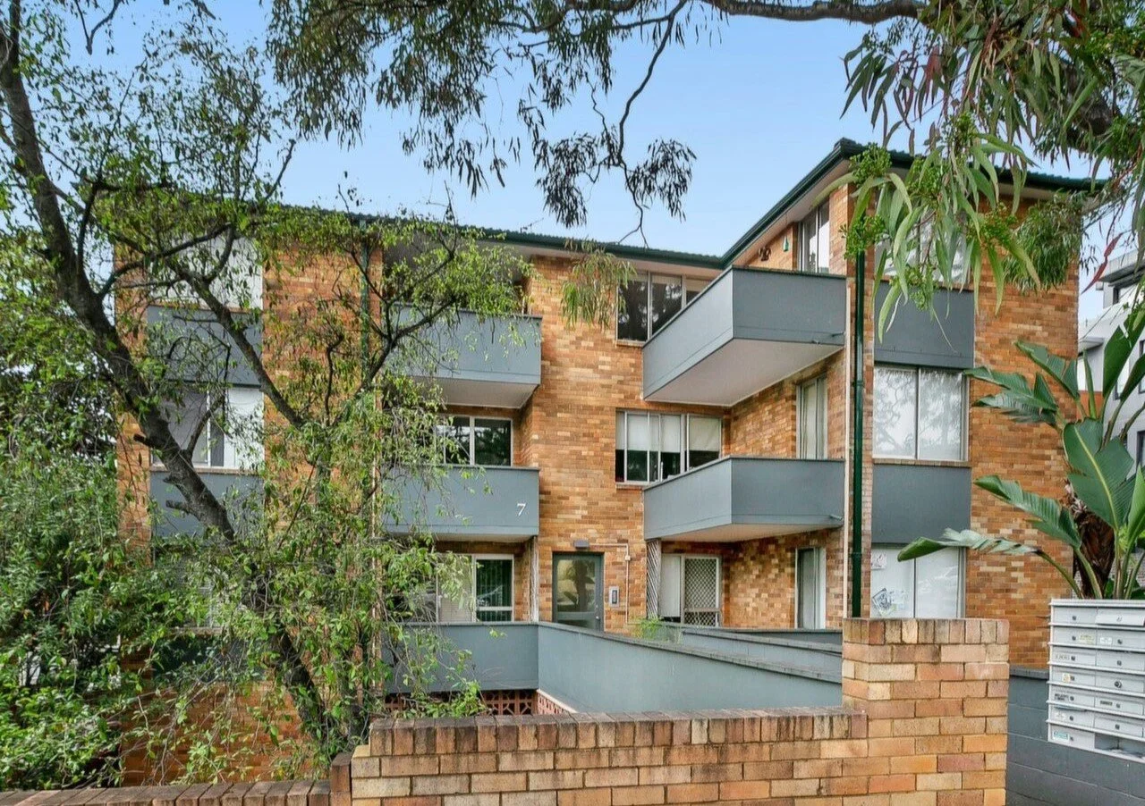 Multi-story brick apartment building with blue balconies surrounded by trees and greenery.