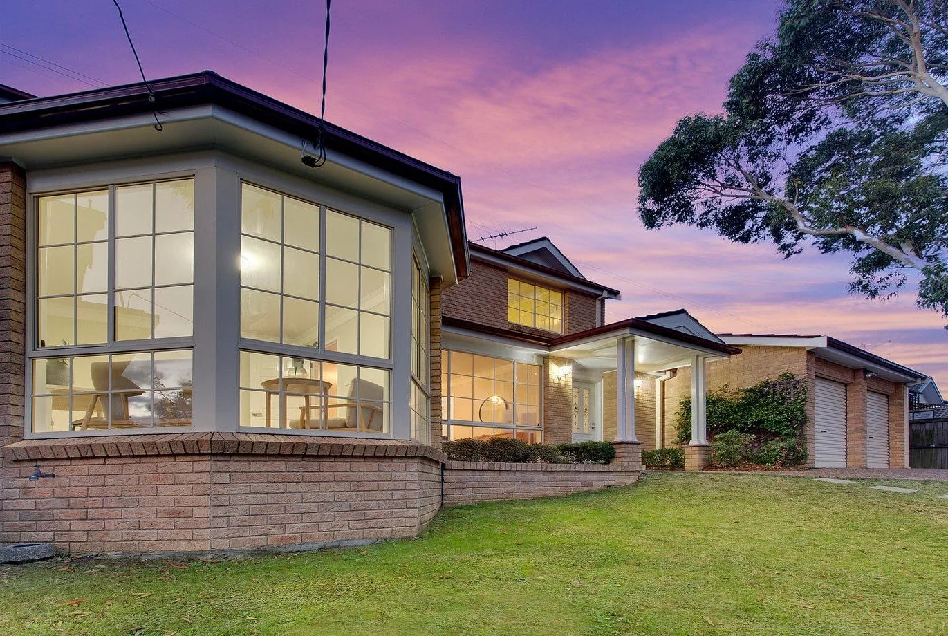 Exterior view of a modern brick house during sunset with large windows, a lawn, and a tree on the right.