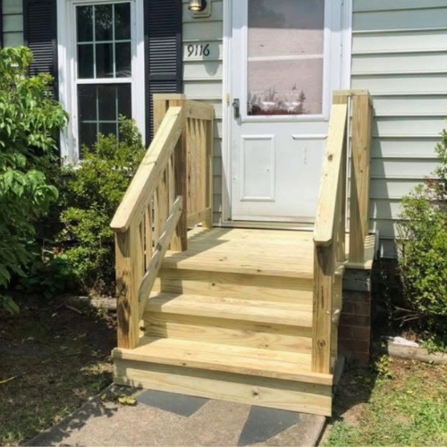 New wooden front porch stairs with handrails installed in front of house with white door and siding, surrounded by shrubs and plants.
