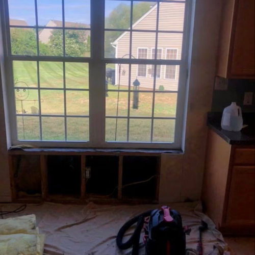 A view of a residential neighborhood through a window inside a house that is undergoing renovations. The window has multiple panes and is partially open. Outside, there is a grassy yard, a neighboring house, and a lamp post. Inside, construction mate