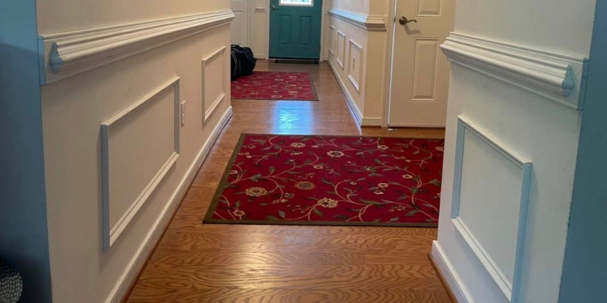 Hallway with hardwood floors, white wainscoting, and red floral area rugs, leading to a teal front door.
