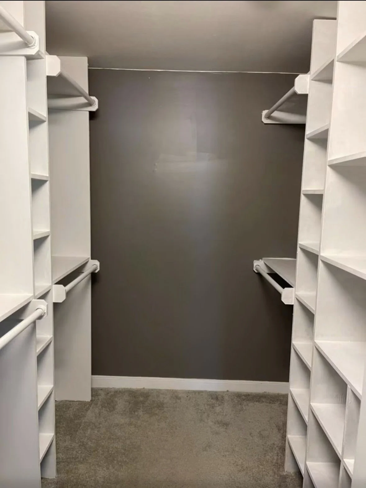 Empty walk-in closet with white shelves and hanging rods on both sides, gray accent wall, beige carpeted floor.