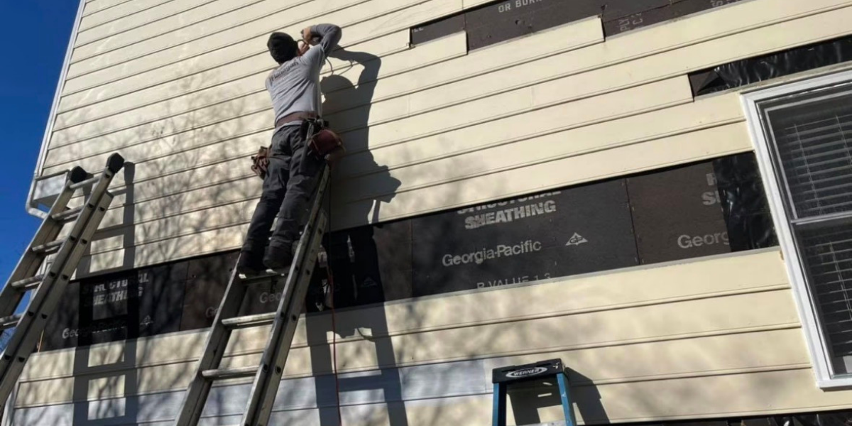 A person working on the exterior wall of a building, standing on a ladder, installing siding panels. There is another ladder on the left side of the building and a small step stool on the ground.