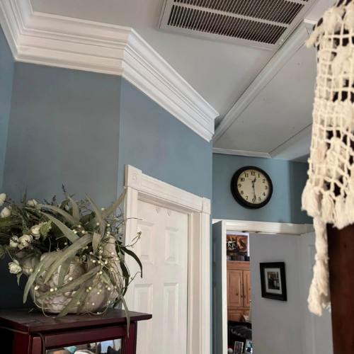 Interior view of a home with blue walls, white crown molding, a white door, a round clock on the wall, and a bouquet of artificial flowers in a vase on a dark wooden table.