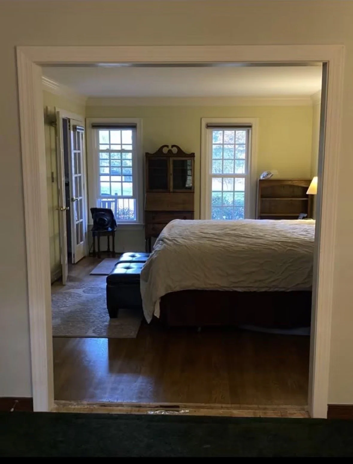 View of a bedroom seen through a doorway, featuring a bed with a white quilt, two windows behind the bed, a wooden dresser with glass doors, and a small wooden bookshelf, with natural light coming in from the windows.