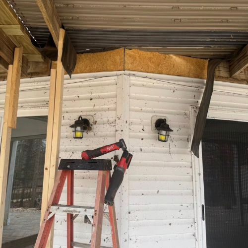 Inside a construction site showing the exterior corner of a building, with a stepladder, two wall-mounted lights, and construction materials like wood and insulation visible.