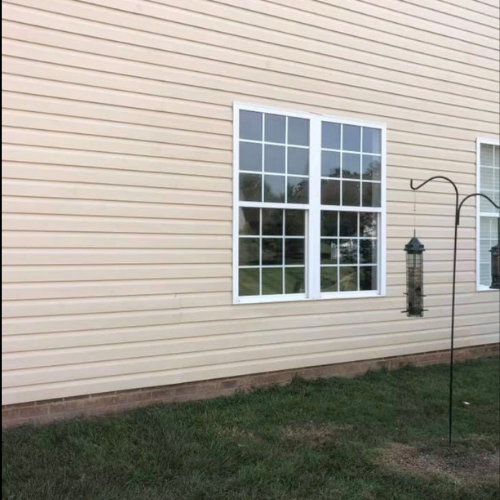 Exterior of a house with light beige vinyl siding, a large window with multiple panes, and a bird feeder hanging from a metal shepherd's hook in the yard.