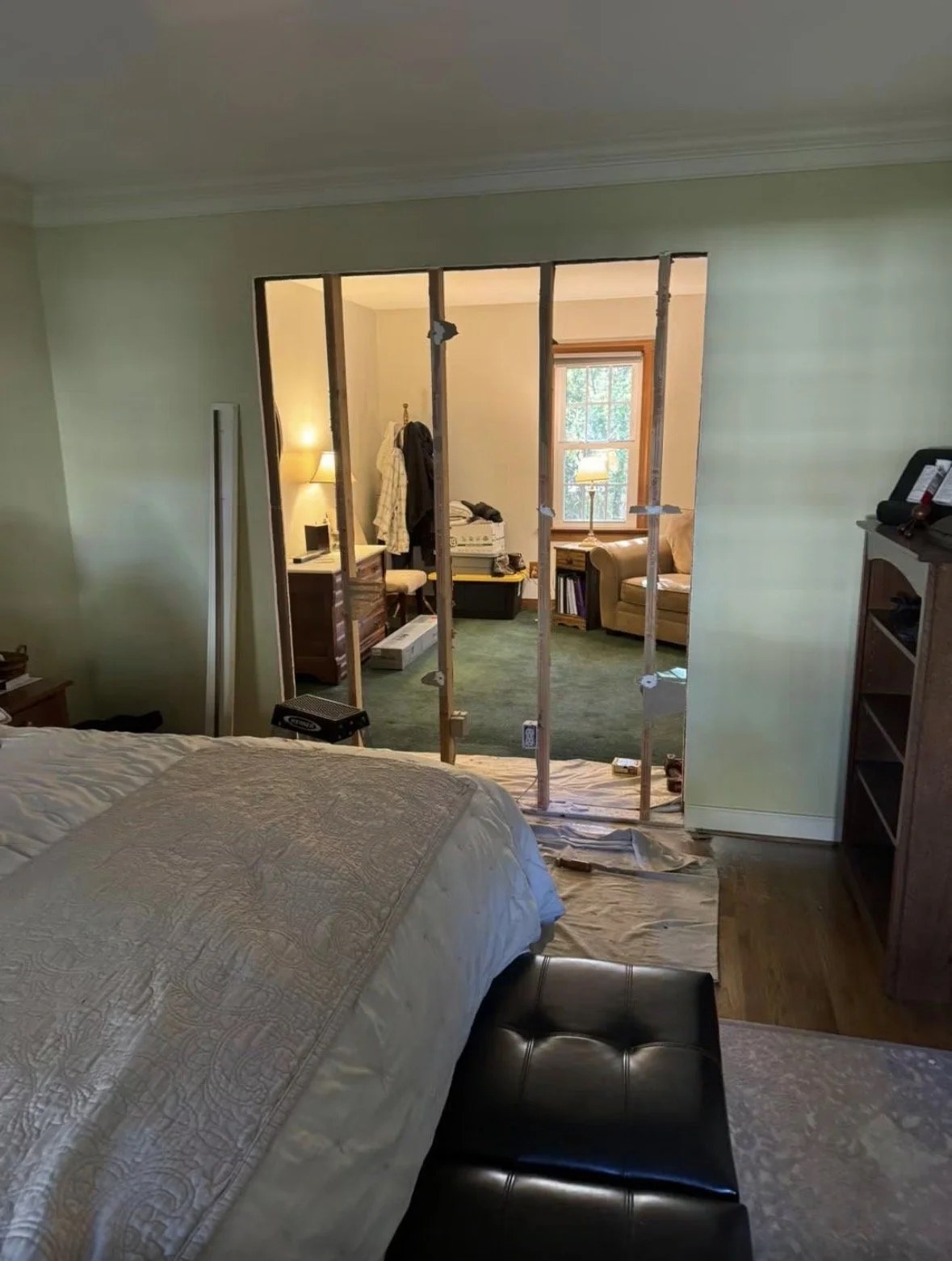 Bedroom with a bed in the foreground and an interior wall removed, showing an adjacent room with furniture and a window.