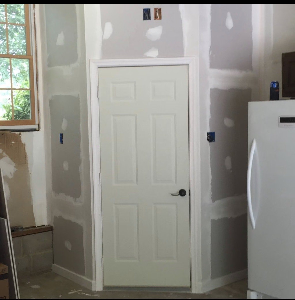 Interior of a room under renovation with unfinished drywall, a closed white door, a window with a wooden frame, and a white refrigerator.
