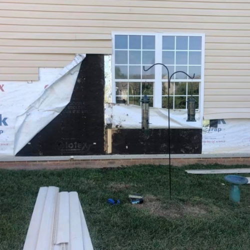 Construction site showing a partially installed exterior wall with an image of a window and a black section, surrounded by construction materials and tools on the grass.