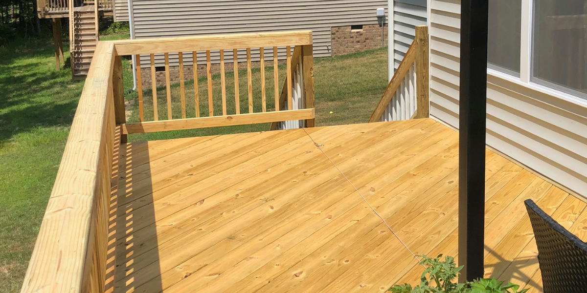 Newly built wooden deck attached to a house with beige siding, featuring a railing on one side and a black support post, with a small section of outdoor furniture visible.
