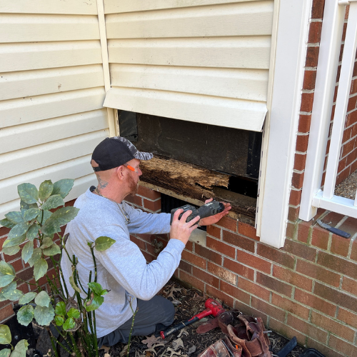 A man wearing a baseball cap, sunglasses, and a light gray shirt is repairing or inspecting a vent or furnace opening on the side of a brick house. He is using a tool to work on the black metal part of the vent, which is partially covered by a white 