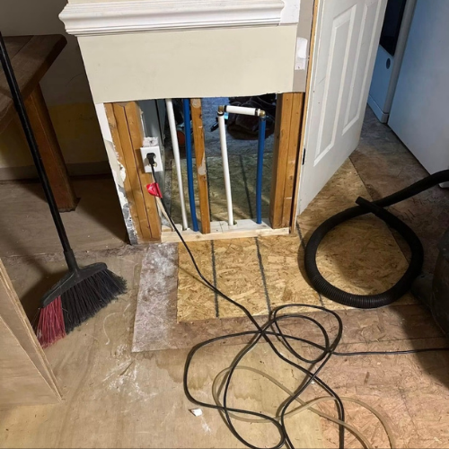 Open wall with electrical wiring, pipes, and a hole for plumbing access. Various cords and a vacuum hose are on the floor.