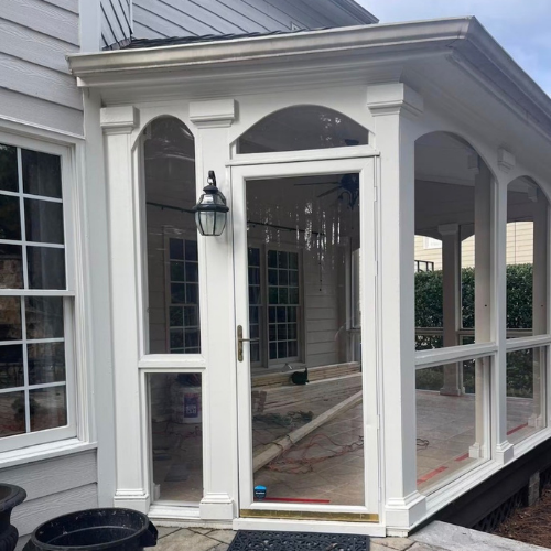 Front view of a small enclosed porch with white framing, glass door, large windows, decorative molding, and an outdoor wall lantern
