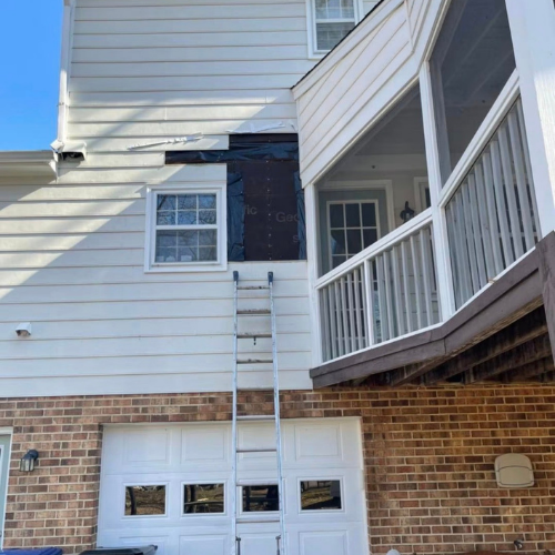 Exterior view of a house with white siding and brick foundation, showing a ladder leaning against the wall and an area with black repair or construction work around a window.