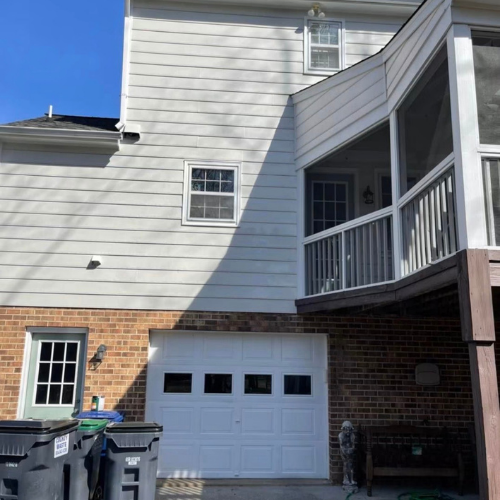 The exterior of a house showing white siding, a brick section, windows, a garage door, and a covered porch with a railing.