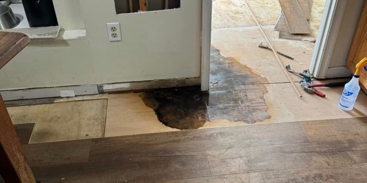 A construction site showing a partially replaced wooden floor with a wet, dark stain near a doorway. Tools and materials are scattered around, including planks, a pry bar, and a spray bottle.