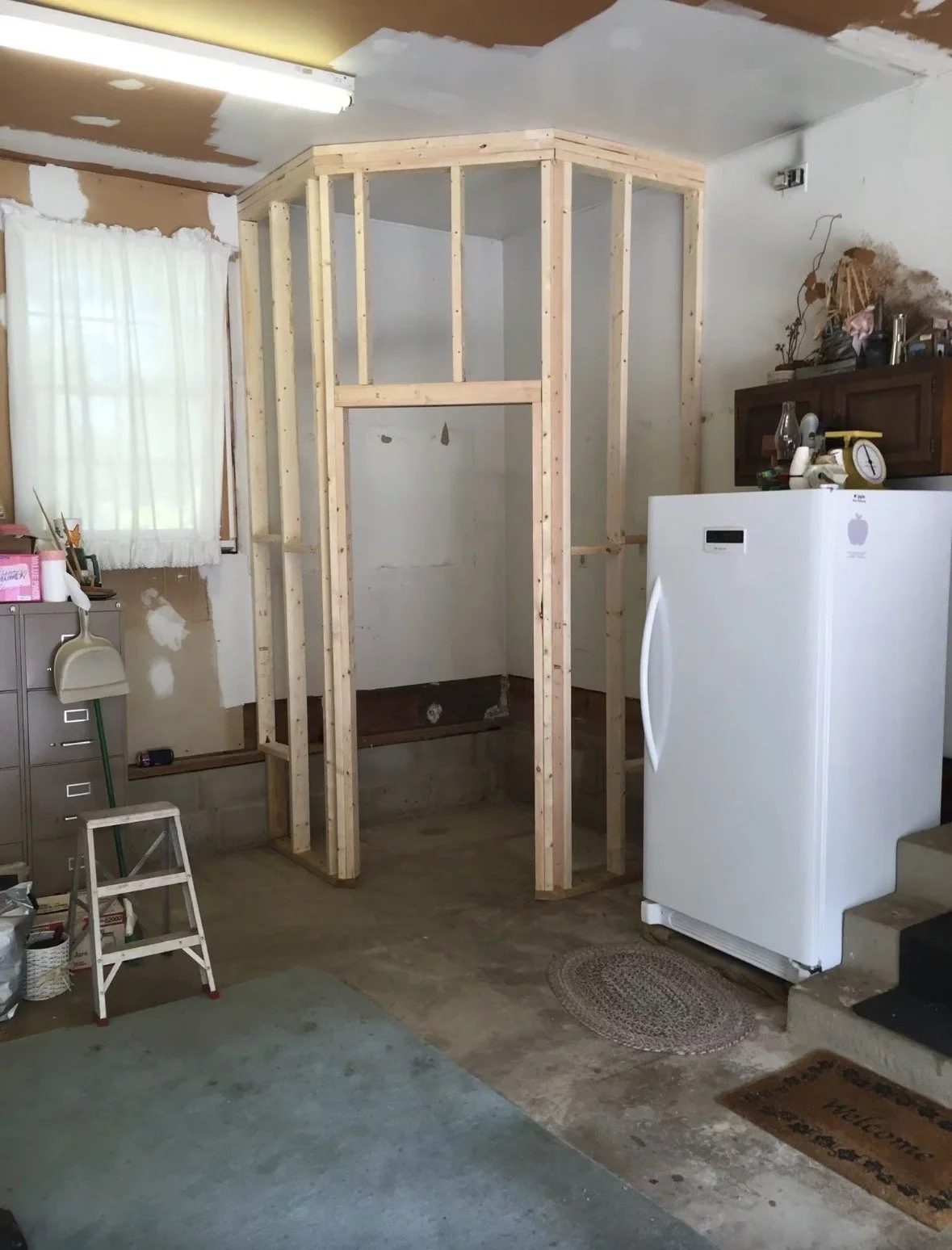 A partially built wooden frame for a shower or small room in a garage with unfinished walls, adjacent to a white refrigerator, a window with a curtain, and various tools and objects.