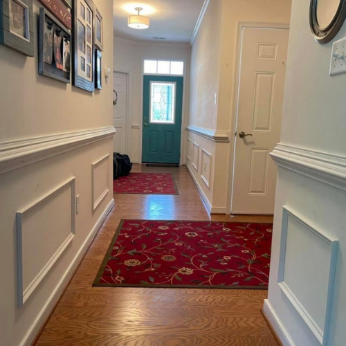 Hallway with wooden floors, white wainscoting, framed photos on the left wall, a red patterned area rug, a teal front door with glass, a hanging ceiling light, and a door on the right side.