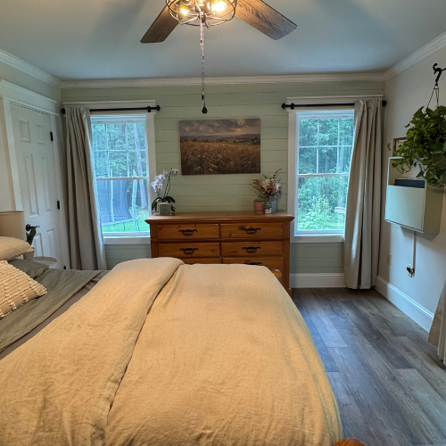 Bedroom with two windows, wooden dresser, framed landscape picture, potted plant, ceiling fan, and a bed with beige bedding.