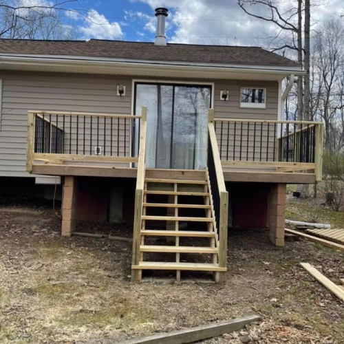 Newly built wooden deck with stairs and metal railings attached to the back of a house, with some construction materials on the ground.