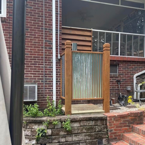 Small outdoor shower or partition with wooden frame and corrugated metal panel, outside a brick building with stairs and a brick patio.