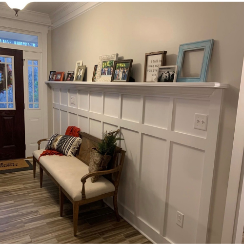 A hallway with a wooden bench, pillows, a potted plant, and a picture frame. A white trim with various framed photos and decorations is displayed on the wall above the trimboard.