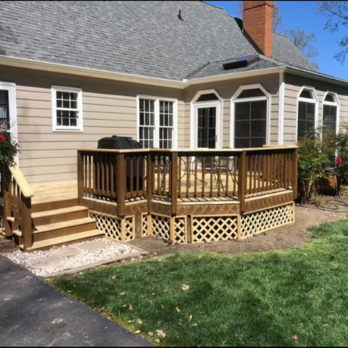 Backyard wooden deck with stairs, enclosed by a railing, attached to a beige house with white-framed windows, surrounded by green grass and plants under a clear blue sky.