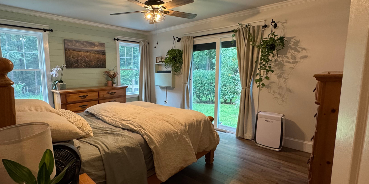 A cozy bedroom with a wooden bed frame and beige bedding, two windows with white curtains and a glass sliding door leading to a green yard, a wooden dresser with decorative items, hanging plants, a wall-mounted heater, and a floor fan near the bed.