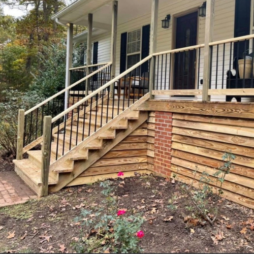 Front porch with stairs leading up to a house, with a garden bed at the base containing pink flowers and a shrub, and a wooded area in the background.