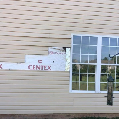Part of a beige house exterior with vinyl siding, a window, and some construction or repair work around the siding.